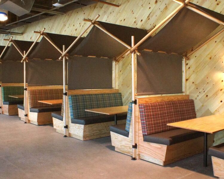 A series of of plywood banquettes or booths with dining tables between them. There are solid seat cushions and plaid back cushions.