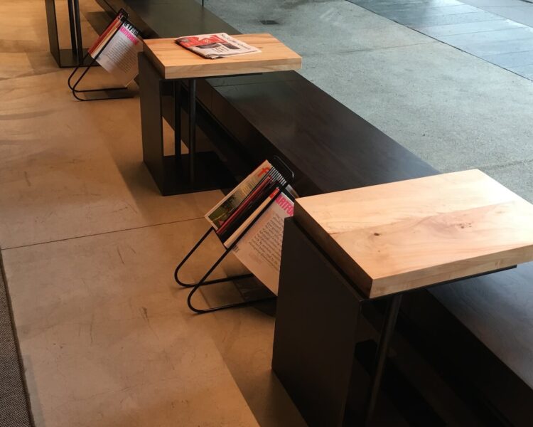 A long, black backless bench in front of a glass wall, with several C-shaped tables tucked underneath it. The tables have light-colored solid wood tops and metal bases.