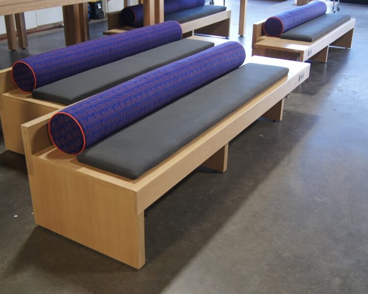 An angled view of a bench in veneer with three support panels. The bench cantilevers beyond the furthest panel. There is power embedded on the front side of the seating platform. There is a long seat cushion and a bolster.